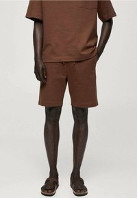 Homme portant une chemise marron à manches courtes et un short assorti avec des poches, associé à des sandales marron, se tenant avec une main dans la poche.