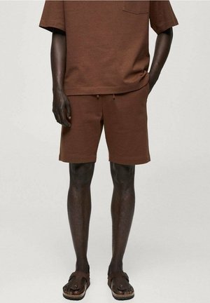 Homme portant une chemise marron à manches courtes et un short assorti avec des poches, associé à des sandales marron, se tenant avec une main dans la poche.
