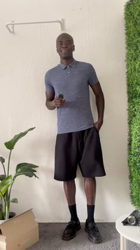Mørkegrå kortermet polo skjorte med teksturert stoff, parret med knelang sorte shorts og sorte sko. Grønn plante og eske nærliggende.
