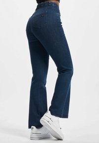 Kék denim, bővülő szárú farmernadrág, magas derékkal, zsebekkel, varrott részletekkel és sima textúrával, fehér sportcipővel párosítva.