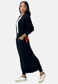 Larga chaqueta negra de punto con textura, de frente abierto y lettering naranja en el bolsillo. Lleva puesta una blusa blanca y pantalones negros, combinada con zapatillas.