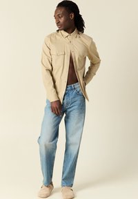 Camicia button-up color tan con due tasche sul petto, abbinata a jeans in denim azzurro chiaro, indossata con scarpe slip-on beige. Linee pulite e vestibilità casual.