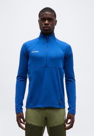 Hombre con un top de manga larga con cremallera cuarteada azul de Mammut y pantalones verde oliva, de pie contra un fondo claro y liso.