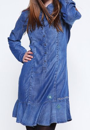 Frau trägt ein blaues Jeanskleid mit langen Ärmeln, Knopfleiste vorne und gerafftem Saum, steht mit einer Hand in der Hüfte und der anderen Hand im Haar.