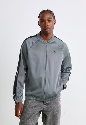 Mand iført grå Jordan zip-up jakke med sorte striber og logo, står med den ene hånd i lommen mod en ensfarvet baggrund.