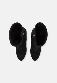 Paire de bottes noires mi-mollet avec des fermetures éclair latérales et des détails subtilement scintillants, vues de dessus sur un fond clair uni.