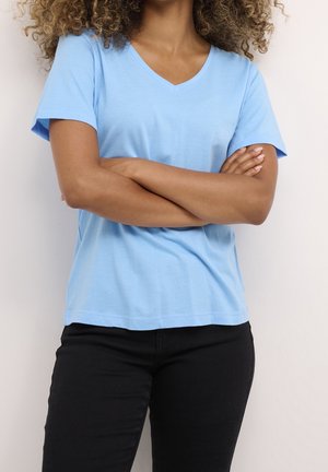 Vrouw met krullend haar, gekleed in een lichtblauw V-hals shirt en zwarte broek, met gekruiste armen tegen een effen witte achtergrond.