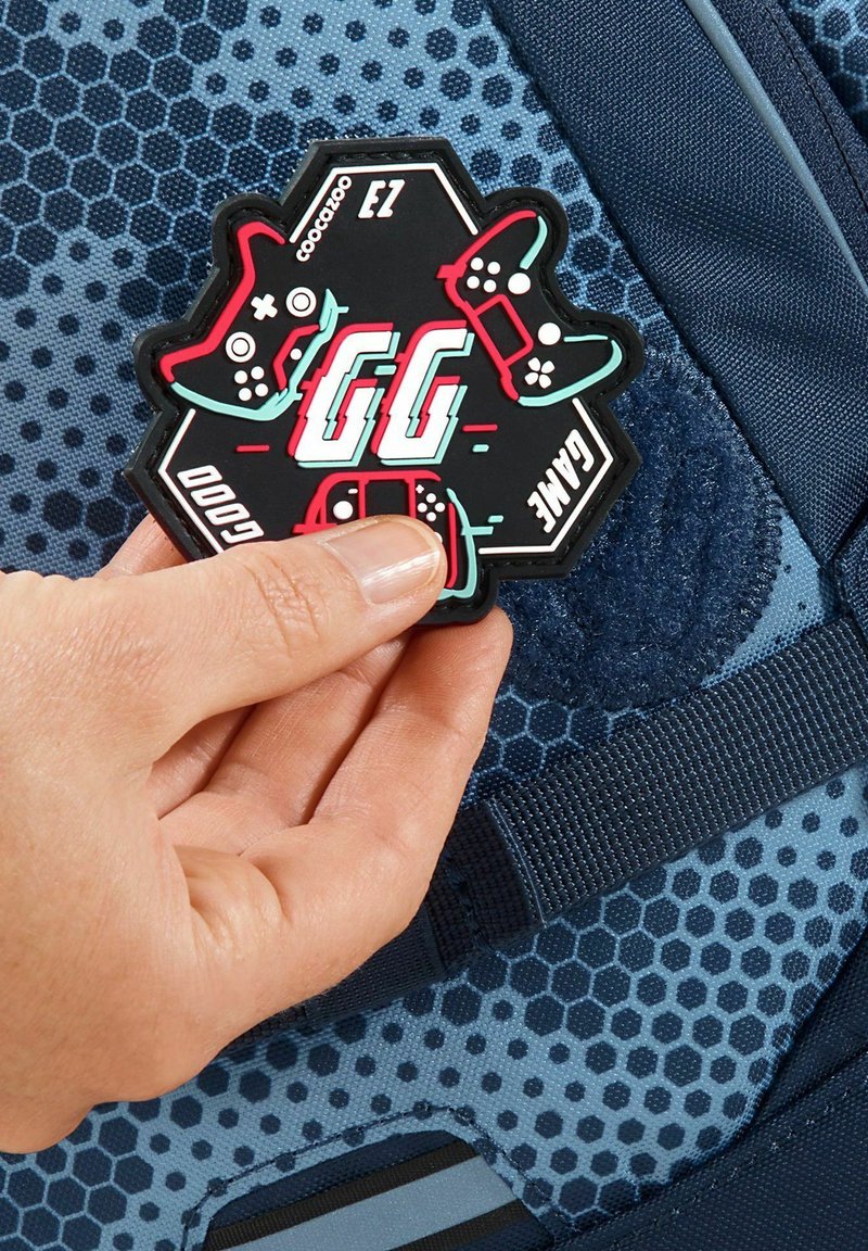 Rubber gaming patch with hexagonal shape, featuring black background, red and teal accents, and game controller graphics. Attached to a blue textured surface.