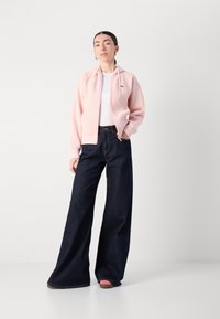 Un sweat à capuche zippé rose clair avec des cordons, porté par-dessus un haut blanc. Assorti avec un jean en denim noir à jambes larges. Les chaussures sont roses claires avec des semelles marron.