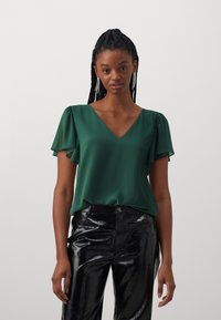Jonge vrouw met gevlochten haar draagt hangende oorbellen, een groene V-hals blouse met fladdermouwen en zwarte glanzende textuurbroek.