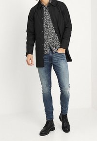 Uomo che indossa un cappotto nero con bottoni, una camicia floreale in bianco e nero, jeans skinny blu leggermente strappati e stivali neri con lacci, in piedi con una mano in tasca.