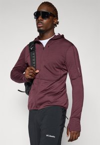 Mørkerød zip-up hættetrøje med hætte, lange ærmer og tommelfingerhuller. Båret med sorte bukser med et Columbia-logo. Sort rygsæk og solbriller.