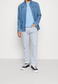 Denimová košile s modrým tričkem pod ní, pruhované světle modré a bílé kalhoty a černé tenisky s bílými podrážkami. Neformální outfit.