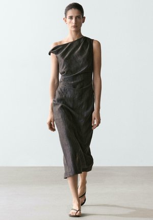 Femme marchant dans une robe charcoal asymétrique sans manches avec des sandales noires à bretelles sur un fond clair uni.