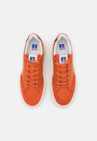 Orange lädersneakers med en slät yta, med kontrasterande vita gummisulor och en märkeslogotyp i blått och rött på tungan.