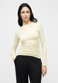 Μπλούζα με μακριά μανίκια σε ανοιχτό κίτρινο χρώμα, κατασκευασμένη από απαλό, ελαστικό ύφασμα με εφαρμοστή γραμμή. Συνδυάζεται με μαύρο παντελόνι. Χωρίς σχέδια ή διακοσμητικά στοιχεία.