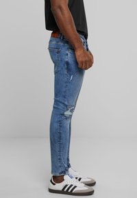 Jeans de mezclilla azul de corte ajustado con detalles desgastados y un parche de cuero marrón en la cintura, combinados con zapatillas blancas y grises.