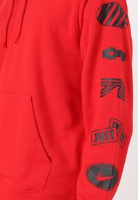 Röd hooded sweatshirt med svarta grafiska tryck på ärmarna, inklusive "JUST DO IT", logotyper och abstrakta mönster. Har en framficka och dragsko på huvan.