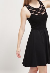 Schwarzes Kleid mit einem Spitzenoberteil und ärmellosem Design. Der weit schwingende Rock reicht unter die Taille und hat eine glatte Textur sowie eine schlichte Silhouette.