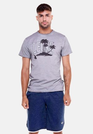 Homme portant un t-shirt gris avec l'inscription "LEONE" et un motif de palmier, associé à un short bleu, debout devant un fond uni clair.