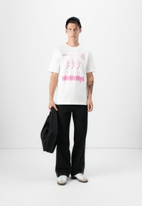 Hvit bomulls T-skjorte med rosa grafisk trykk og "adidas"-logo. Sammen med svarte baggy bukser og hvite sneakers.