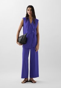 Blazer senza maniche viola con un solo bottone, abbinato a pantaloni viola a gamba larga. Il modello tiene una borsa a tracolla nera con texture.