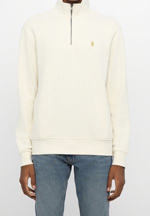 Sudadera blanca con cremallera hasta un cuarto, con un pequeño logo bordado en amarillo en el lado izquierdo del pecho, combinada con vaqueros azules.