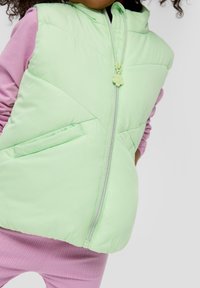 Helles grünes, pufferartiges Weste mit einem Reißverschluss, ausgestattet mit zwei Seitentaschen und einem blumenförmigen Reißverschluss-Anhänger, getragen über einem pinken Langarmoberteil.
