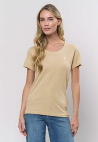 Béžové tričko s krátkým rukávem z měkké bavlny, s kulatým výstřihem a malým logem na levé hrudi.