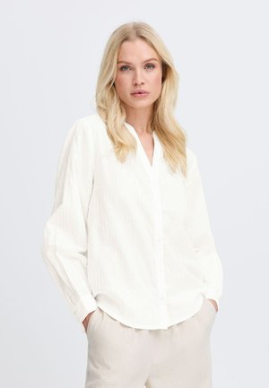 Chemise blanche à manches longues bouffantes avec un motif à rayures verticales et un col en V, associée à un pantalon beige clair.