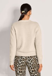 Frau mit dunklen Haaren im Pferdeschwanz, die einen beigen Strickpullover und Hosen mit Leopardenmuster trägt, von hinten vor einem neutralen Hintergrund gezeigt.