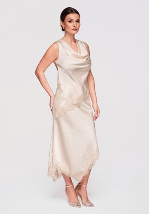 Kobieta stojąca w bezrękawnikowej, satynowej sukience midi w kolorze szampańskim z koronkową lamówką oraz w wysokich butach na obcasie na białym tle.
