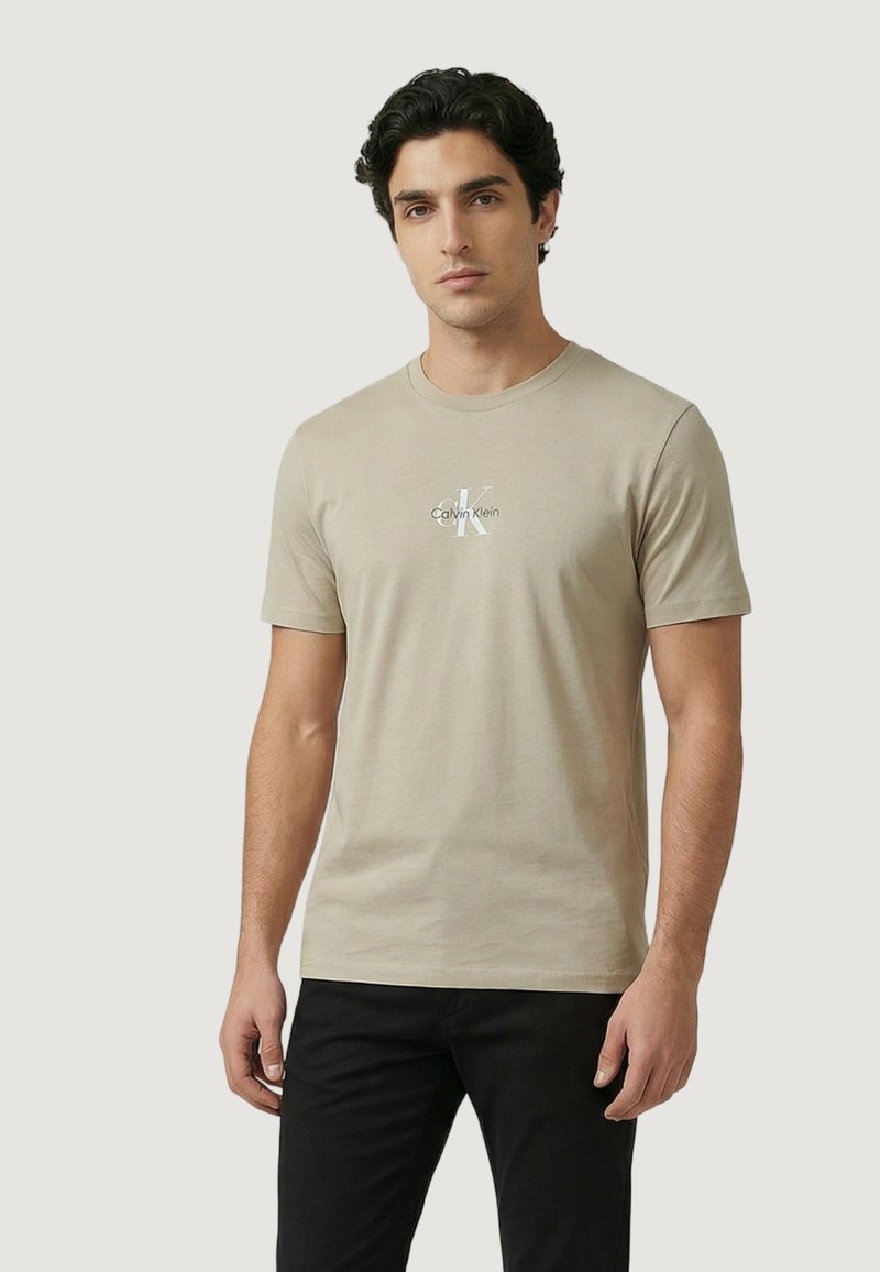 Homme portant un t-shirt à manches courtes beige Calvin Klein et un pantalon noir, debout devant un fond clair uni.