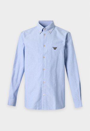 Homme debout portant une chemise bleu clair boutonnée, un pantalon plissé beige, une ceinture noire, des chaussures noires et un collier de perles sur fond uni.
