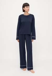 Pajama set u tamnoplavoj boji od mekog materijala; dugih rukava, s bijelim rubom, opuštenog kroja, uparen s odgovarajućim dugim hlačama koje također imaju bijele detalje.