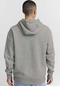 Sudadera gris con tejido texturizado. Presenta una capucha con cordón y puños y dobladillo acanalados. Vista trasera, combinada con jeans de color azul claro.