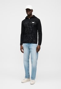 Μαύρο μπουφάν με κουκούλα με καμουφλάζ σχέδιο, κλείσιμο με φερμουάρ και λογότυπο, σε συνδυασμό με ανοιχτό μπλε τζιν και λευκά αθλητικά παπούτσια.