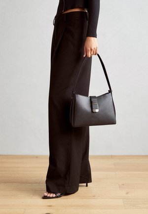 Kobieta ubrana w czarne spodnie z szerokimi nogawkami i sandały na obcasie, trzymająca czarną skórzaną torebkę z klamrą.