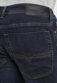 Vaqueros de mezclilla en azul oscuro con costuras doradas visibles. Presentan un parche de cuero de marca y un bolsillo trasero con un diseño distintivo en forma de V.