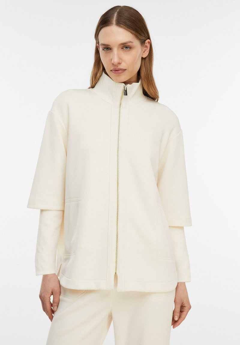 Femme portant une veste zippée crème à manches courtes sur manches longues et un pantalon crème assorti, sur un fond uni.