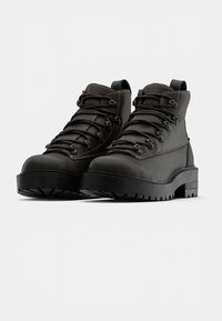 Hnědé kožené turistické boty s černými gumovými podrážkami, kulatými tkaničkami, kovovými průvleky a polstrovaným límcem u kotníku.