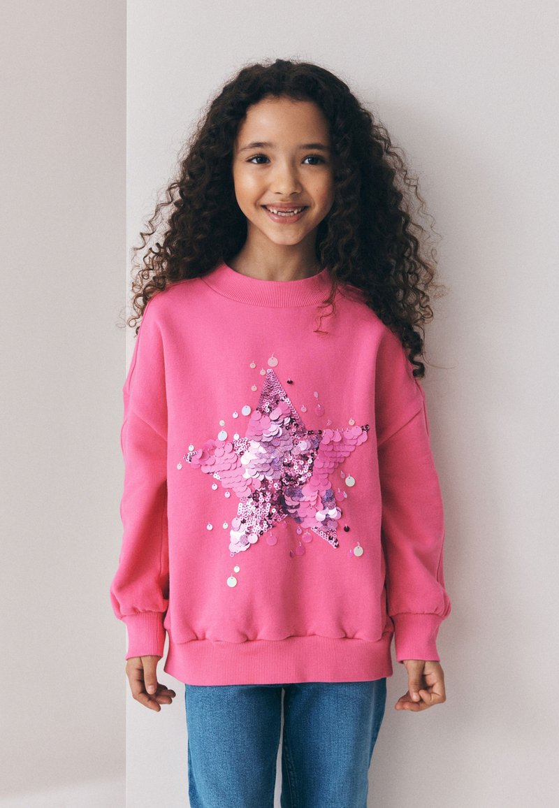 Niña sonriente con cabello largo y rizado, que lleva un suéter rosa brillante con una gran estrella de lentejuelas y vaqueros azules, de pie frente a una pared lisa.