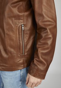 Braune Lederjacke mit glatter Oberfläche, ausgestattet mit einer seitlichen Reißverschlusstasche und taillierter Passform. Die Ärmel sind leicht angepasst.