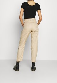 Beige cotton trousers with a tapered cut. Features two back pockets and a label on the right pocket. Worn with black ankle boots.
