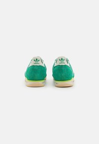 Zapatillas de ante verdes con un perfil bajo, que presentan una suela y un talón de color crema, y un logo verde en la parte posterior.