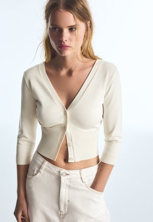 Junge Frau mit blondem Haar trägt eine cremefarbene, kurze Strickjacke und hellfarbene, hoch taillierte Hosen vor einem schlichten weißen Hintergrund.