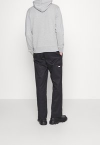 Vista posteriore di una persona che indossa un felpa grigia chiara, pantaloni da lavoro neri con una toppa con logo e scarpe nere con suola spessa, in piedi contro uno sfondo neutro.