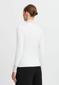 Hvid langærmet top med en høj blondekrave og blondeaccenter ved manchetterne. Flared design med en glat, ribbet struktur.