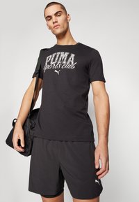 Camiseta de algodón negra con gráfico en blanco "PUMA sports club", combinada con pantalones cortos negros. El modelo sostiene una bolsa de deporte negra.