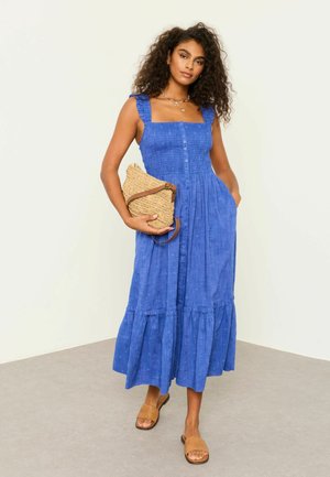 Frau mit lockigem Haar trägt ein blaues ärmelloses knöpfbares Kleid, trägt eine gewebte Clutch und trägt braune gewebte Slip-On-Sandalen.